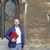 English professor with beard holds red book outside a stone building