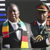 A man stands behind Emmerson Mnangagwa while he speaks at an event.