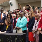Dozens of people stand around Gretchen Whitmer as she signs legislation while sitting behind a desk outside.