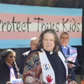 Pat Langlois speaks on stage, with several people holding transgender pride flags behind her.