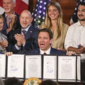 Ron DeSantis holds up signed documents as several people applaud while standing behind him.