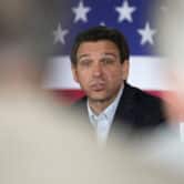 Ron DeSantis speaks at an event, with a large American flag in the background.