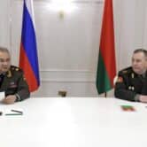 Sergei Shoigu and Viktor Khrenin sit at a white table while speaking to the media, with a Russian flag and a Belarusian flag in the background.