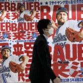An Asian man wearing a white face mask walks past posters of Trevor Bauer throwing a baseball while wearing a Yokohoma DeNA BayStars uniform.