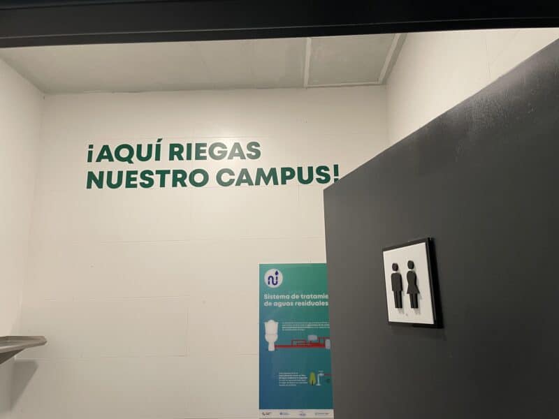 A black door on the right has the man/woman bathroom sign on it. Text and an infographic on the wall to the left in Spanish tells users that the water they use here will be treated and used to water plants on the university campus.