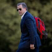 Hunter Biden walks while wearing aviator sunglasses, a blue suit and a red backpack.