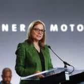 Marry Barra speaks on stage during an event, with a few people seated behind her and "General Motors" displayed on a screen.