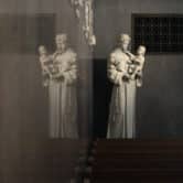 A statue of St. Anthony holding the infant Jesus is reflected against a wall behind several rows of pews.