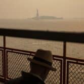 A person sits on the State Island Ferry with the Statue of Liberty slightly obscured by a haze-filled sky in the background.
