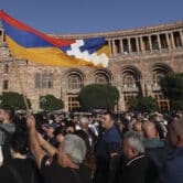 A crowd of people are protesting outside a government building in Armenia, one person is flying the flag of the Republic of Artsakh