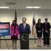 California Attorney General Rob Bonta speaks at a podium, three women are standing behind him at a press conference.