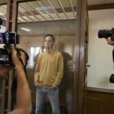 Evan Gershkovich stands behind a glass cage as journalists points their cameras at him.