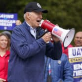 Joe Biden speaks into a megaphone in front of striking United Auto Workers holding signs.