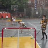 Seven children play ball hockey on a basketball court on a rainy day, with an adult holding a hockey stick looking on.