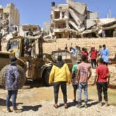 A group of people stand amid destroyed buildings in Libya, among them is a tractor ridden by uniformed service members