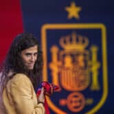 Montse Tome holds a Spanish soccer jersey, with the coat of arms of the king of Spain on a wall in the background.