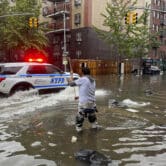 An NYPD SUV drives through flooding in New York City, a man in the foreground is standing in knee-deep water attempting to clear a drain using a long stick.