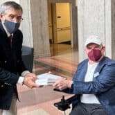 A man in a wheelchair hands a brooklet with more than one hundred pages to a man standing up, with both of them wearing face masks.