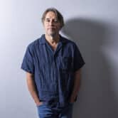 Richard Linklater stands in front of a gray wall while posing for a portrait.