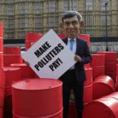 A person wears a fake head modeled after Rishi Sunak during a protest with more than a dozen red barrels.