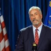 A man in a suit stands behind a microphone, with the flags of the United States and Nevada behind him.