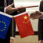 A person hands another person a folder, with miniature European Union and Chinese flags in front of them.