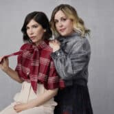 Carrie Brownstein and Corin Tucker poses for a photo in front of a gray wall.