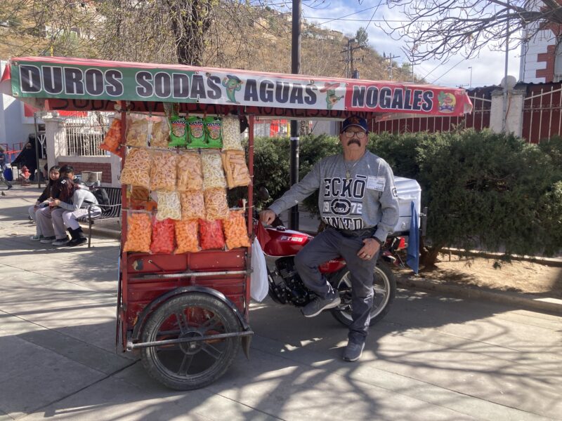 A man with a hat and black mustache sits on his motorbike attached to a snack cart.