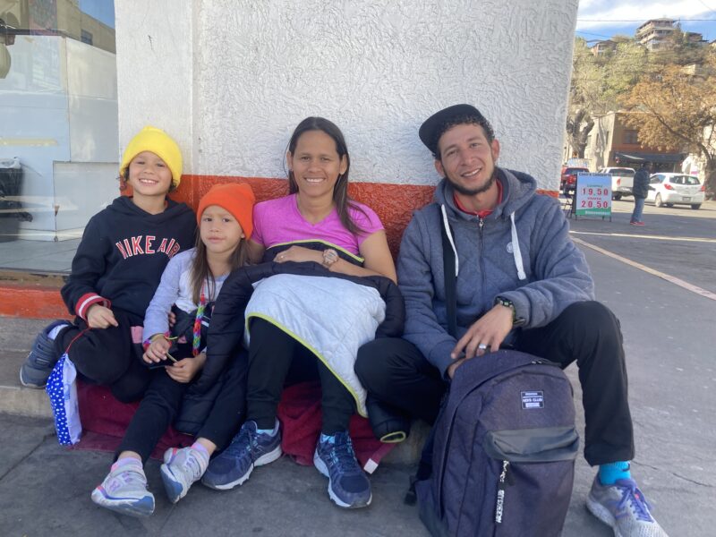 Two adults and their two children sit on a street corner in front of a white painted building in Mexico.