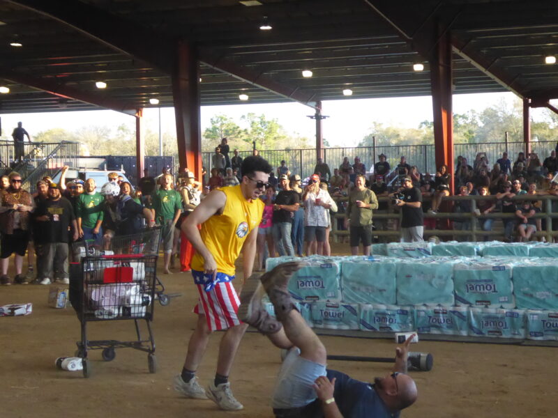 A man in American flag shorts and yellow shirt knocks his rival down while grabbing toilet paper in a cart