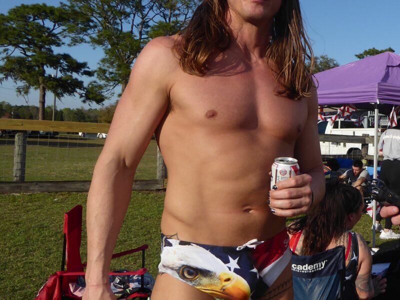 A long-haired shirtless white man wearing mirrored sunglasses and a small underwear-type shorts with a bald eagle and American flag printed on them holds a beer as he stands in front of some camp chairs in a field in front of a tree.