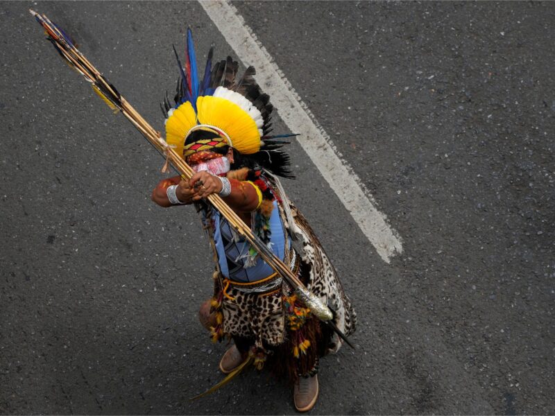 A man wearing Indigenous clothing and face paint points his bow and arrow in the sky while standing on a road.