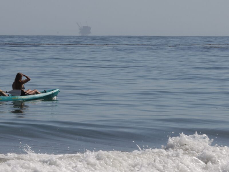 Two people on a green kayak look out towards a oil rig in the distance