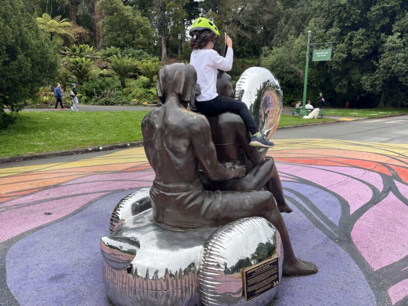 A child wearing a helmet sits on a bronze sculpture of two figures in swim trunks sitting back-to-back on metallic inflatable rings, part of an outdoor art installation