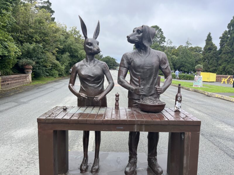 Bronze sculpture of two humanoid figures with animal heads-one a rabbit and the other a dog-standing behind a table.