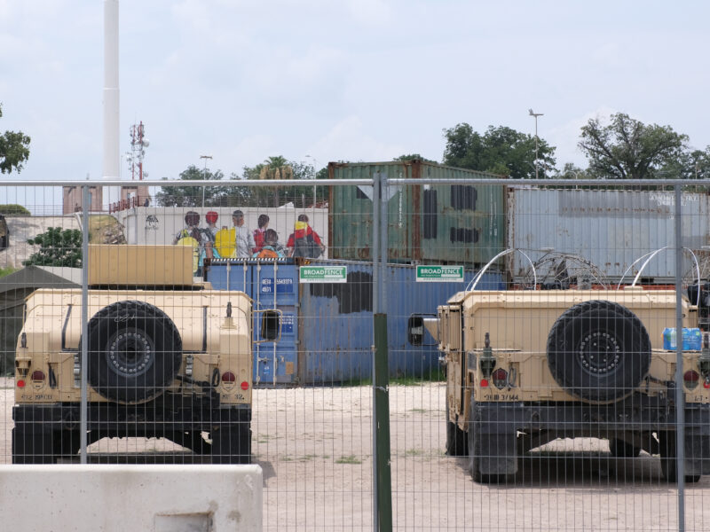 Through a fence, two military vehicles sit vacant with a stack of shipping containers behind them.