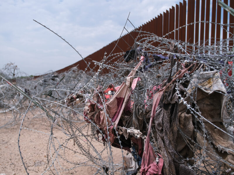 Next to a large rust-colored fence, piles of circular razor wire sit with tattered clothes trapped in the sharp blades.
