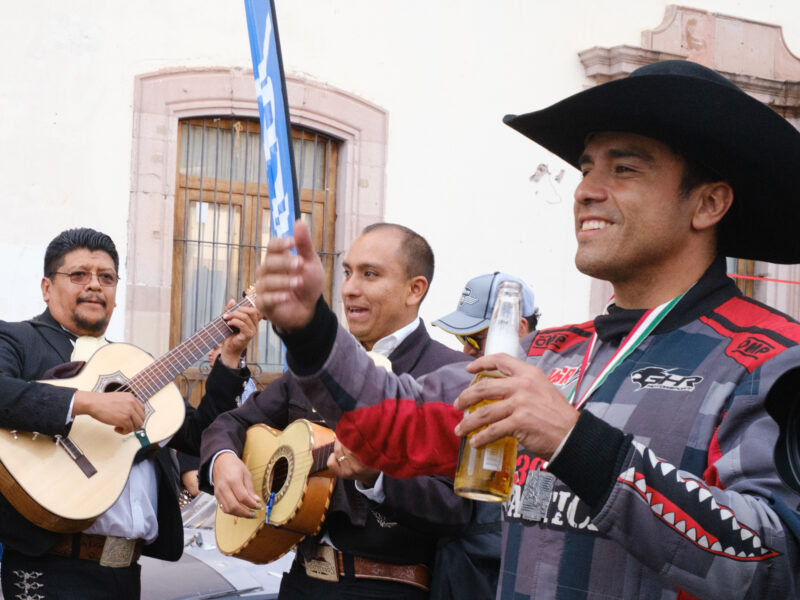 A man with a medal around his neck a black cowboy hat drinks a beer with mariachis behind him.