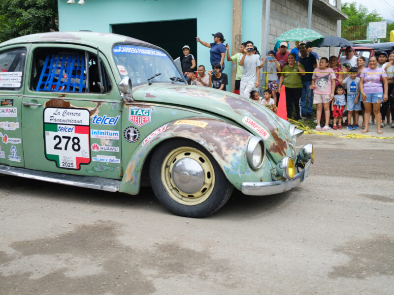 Onlookers take pictures with their cell phones and smile as a Volkswagen drives by on a semi dirt road in Mexico.