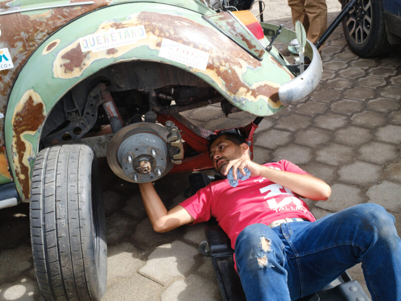 A man in a red shirt works under a patina-green VW Beetle.