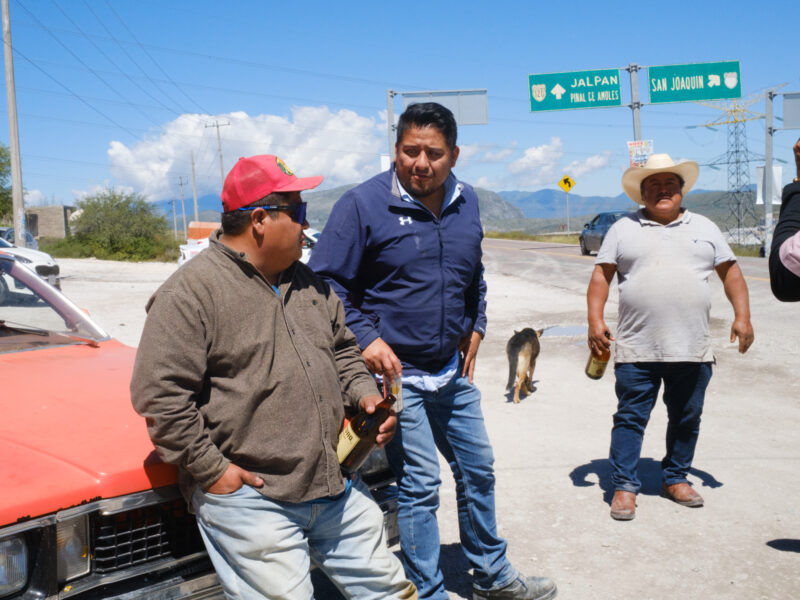 Three men stand by a rural road in Mexico one leans against a red car drinking beer.
