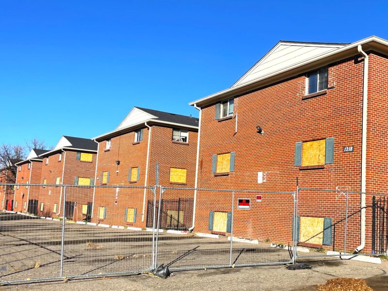 Boarded-up red brick apartment buildings surrounded by a chain-link security fence under a clear blue sky.