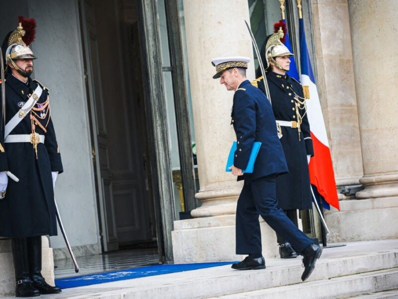 A man in a military uniform walks between soldiers in dress uniform with swords into an open doorway.
