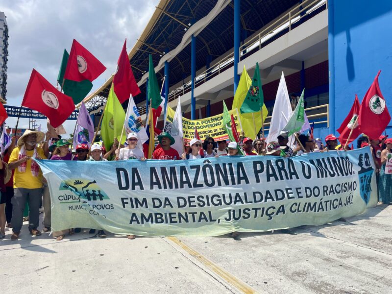 Crowd holds a long banner calling for climate justice, surrounded by colorful flags and signs during a march in Belem.