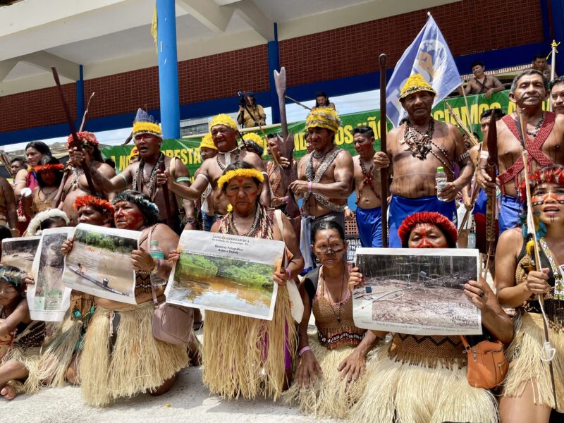 Group of Indigenous leaders wearing traditional attire hold large printed photos showing environmental destruction, during the Great People's March in Belem.
