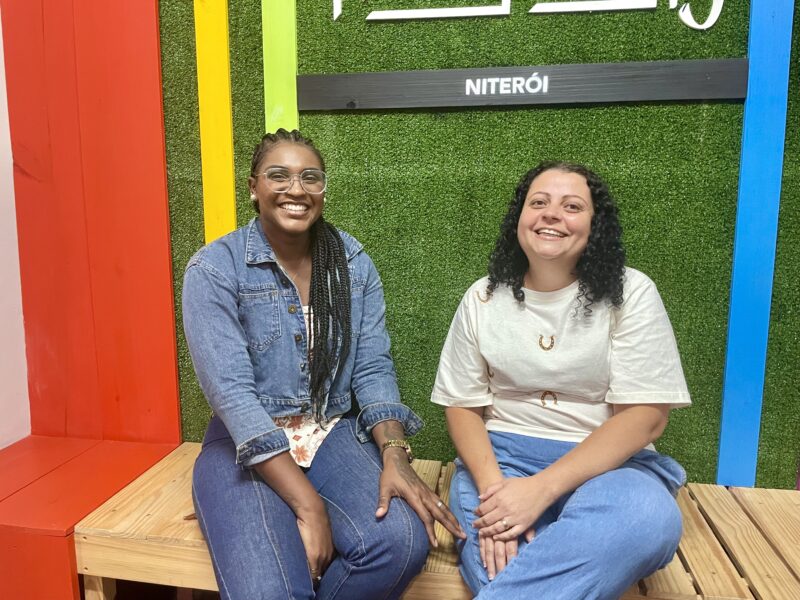 Mariane de Oliveira and Ana Paula Cunha, beneficiaries of Niteroi's Arariboia program, seated and smiling at a municipal building.