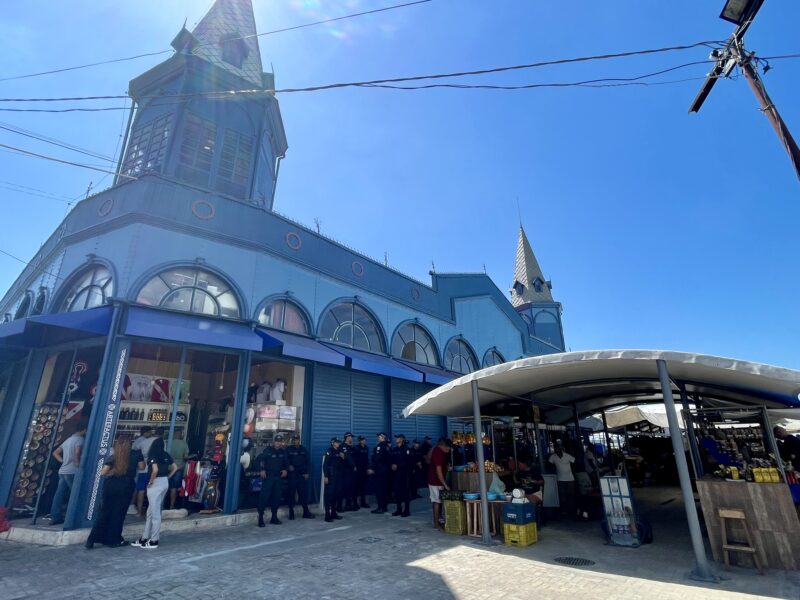 Exterior view of the Ver-o-Peso market in Belem, Brazil, showing vendors and police officers near the entrance under a clear blue sky.