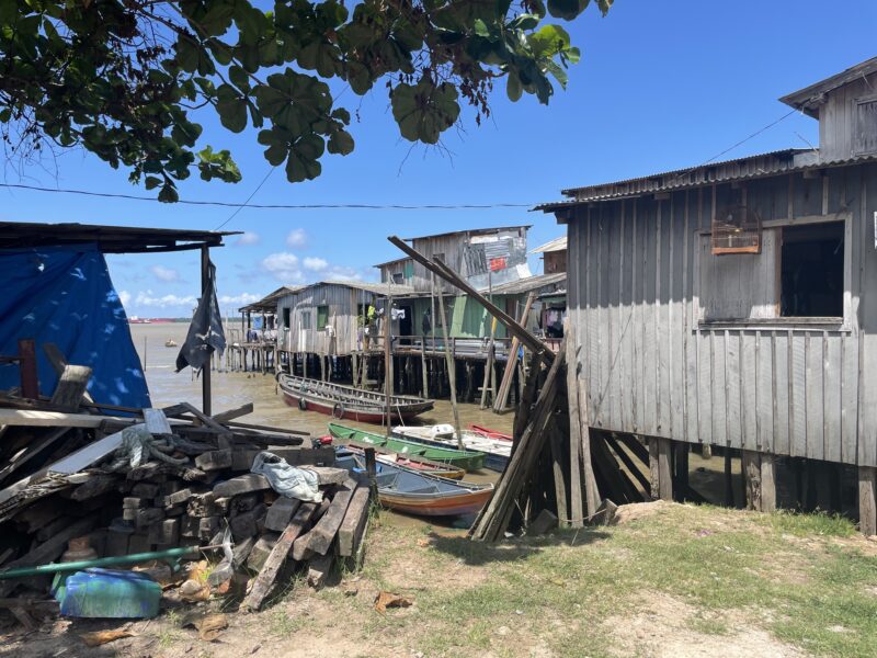 Wooden stilt houses line the river in Vila da Barca, Belem, with small boats moored along the shore and piles of construction materials in the foreground.