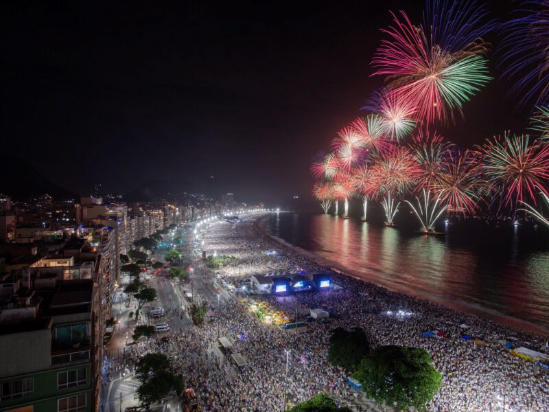 Aerial view of Copacabana beach packed with crowds dressed mostly in white as fireworks explode over the ocean during New Year's Eve in Rio de Janeiro.