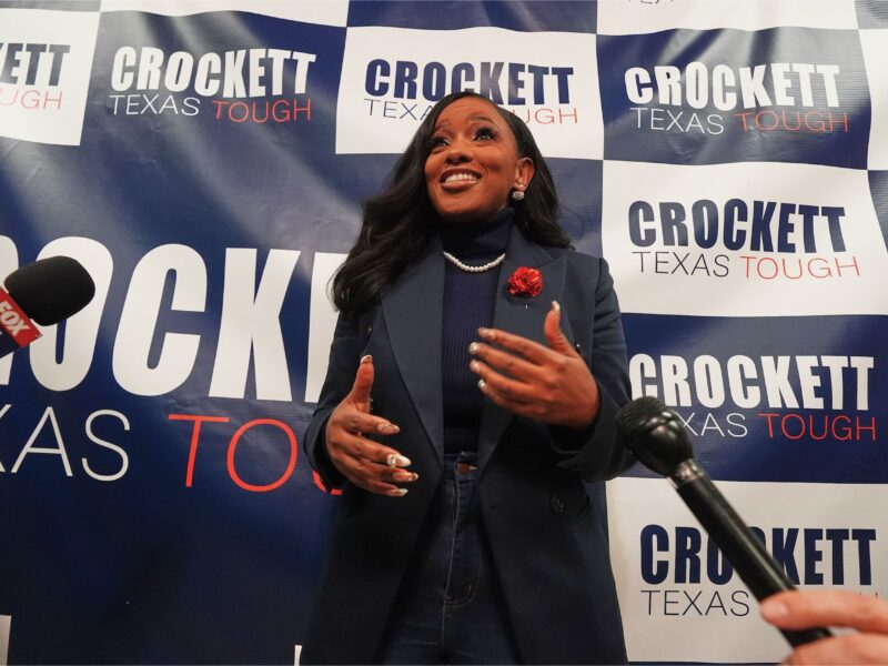 Rep. Jasmine Crockett speaks enthusiastically to reporters while standing in front of a campaign backdrop that reads 